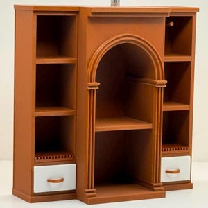 Op de afbeelding: Een bruine miniatuurkast met een centrale boogvormige nis, geflankeerd door twee zijsecties met open planken. Elke zijsectie bevat onderaan een kleine witte lade met een bruine handgreep. De structuur heeft een gladde afwerking.