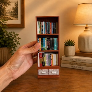 May include: A miniature brown wooden bookshelf featuring three shelves filled with tiny, colorful books and two small drawers at the bottom with a white geometric pattern. The unit is held in a hand, displaying a collection of various book spines.