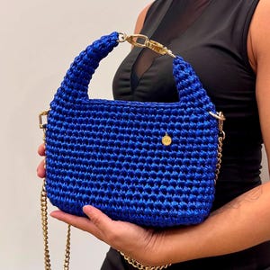 May include: A vibrant royal blue crochet handbag featuring a structured rectangular body, a thick braided handle, and a gold-toned metal chain shoulder strap. A small circular gold charm is attached to the front, and gold hardware connects the handle.