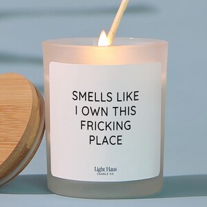 May include: A lit candle in a frosted glass jar with a wooden lid. The label reads "Smells Like I Own This Fricking Place" with "Light Haus Candle Co." below. A matchstick is lit and held above the candle.