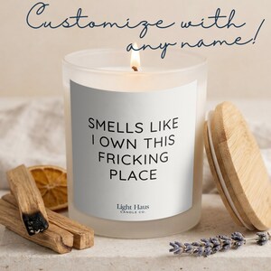 May include: A lit white candle with the text "SMELLS LIKE I OWN THIS FRICKING PLACE" on a white label. The candle is in a frosted glass jar with a wooden lid. Dried lavender, wood sticks, and a dried orange slice are next to the candle.