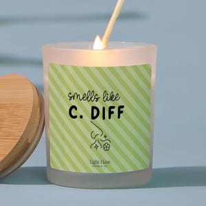 May include: A lit candle in a frosted glass jar with a wooden lid. The label reads "smells like C. DIFF" with a graphic of a nose and flowers. A match is lit above the candle. The candle is from Light Haus Candle Co.