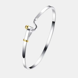 May include: A silver-toned bangle bracelet featuring a hook-and-loop closure mechanism. The design includes a small, polished gold-toned sphere at the end of the hook and two thin gold-toned bands encircling the lower portion of the silver loop.