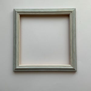 May include: Square picture frame with a light blue and grey wood grain finish. The frame has a simple, classic design, ideal for displaying photos or artwork. The frame is made of wood and has a neutral colour palette.