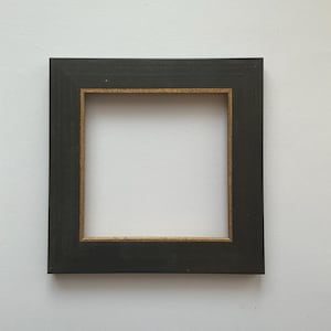 May include: A square picture frame with a matte black finish on the outer border and a thin, metallic gold inner edge. The frame features a flat, wide profile with clean, straight lines and sharp corners, creating a simple and structured appearance.