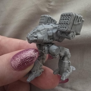 Puede incluir: Un robot en miniatura gris impreso en 3D con un diseño detallado. El robot tiene una cabeza cuadrada, dos lanzadores de misiles en la espalda y piernas articuladas. La miniatura se sostiene en una mano, mostrando su tamaño y características.