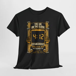Puede incluir: Camiseta negra con un gráfico dorado. El diseño incluye el texto "Yinz Are On The Clock" y "4:12". Debajo, se lee "Draft Weekend April 23-25, 2026 Pittsburgh, PA". El gráfico presenta puentes y un horizonte.