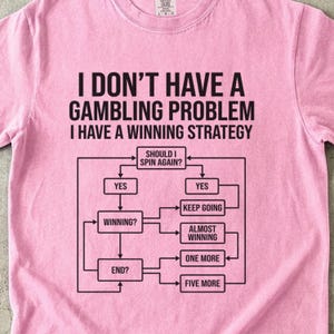 May include: A light pink t-shirt with the text "I DON'T HAVE A GAMBLING PROBLEM I HAVE A WINNING STRATEGY" in black. Below the text is a flow chart with the question "SHOULD I SPIN AGAIN?"
