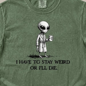 May include: Green Comfort Colors t-shirt featuring a black and white illustration of an alien in a robe holding a cup. The text "I HAVE TO STAY WEIRD OR I'LL DIE" is printed below the alien graphic. The shirt has a crew neck.