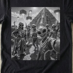 Op de afbeelding: Een zwart t-shirt met een grijstinten grafische print van verschillende buitenaardse wezens in futuristisch harnas die een selfie maken voor een grote stenen piramidestructuur met zwevende geometrische blokken in de lucht boven de scène.