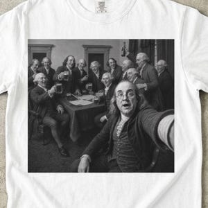 Puede incluir: Una camiseta blanca con un estampado gráfico en blanco y negro de figuras históricas con vestimenta del siglo XVIII reunidas alrededor de una mesa con jarras de cerveza, con una figura en primer plano tomándose un selfie. Etiqueta Comfort Colors.