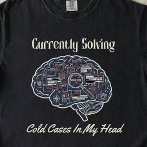 May include: Dark gray t-shirt with the text "Currently Solving Cold Cases In My Head." A graphic of a brain with detective case files and a magnifying glass is in the center.