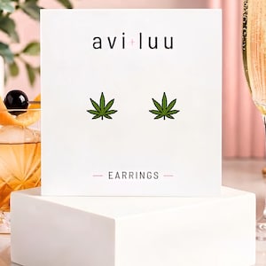 May include: A pair of stud earrings featuring green cannabis leaf designs, displayed on a white square card with the text avi+luu and EARRINGS printed in black. The earrings are positioned centrally on the card, which rests on a white block.