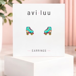 May include: A pair of stud earrings shaped like roller skates on a white display card. The skates are teal with yellow laces, pink wheels, and a small yellow lightning bolt detail. The card features the text avi+luu and EARRINGS.