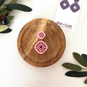 May include: A wooden tray holds two pink and red clay earring cutters and a pair of purple polymer clay earrings. The earrings are square with a floral design and are displayed on a white card with the text "Avi + Luu".