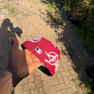 Puede incluir: Una gorra de béisbol roja con letras blancas bordadas en cursiva en el lateral y un pequeño parche con el logotipo de la Major League Baseball. La gorra tiene una corona estructurada y la parte inferior de la visera en color negro.