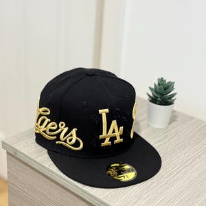 以下が含まれることがあります： 前面にDodgersの文字とLAのロゴがゴールドで刺繍された黒いベースボールキャップ。キャップにはフラットなつばがあり、サイズ7 5/8を示す円形のステッカーが貼られている。側面にもゴールドの刺繍が施されている。