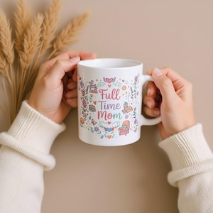 以下が含まれることがあります： ピンクと紫の文字で「Full Time Mom」と書かれた白いセラミックマグ。カラフルな花とハートのイラストで囲まれています。マグには白いハンドルがあり、白いセーターを着た人が持っています。