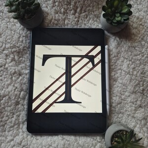May include: A black tablet displays a design featuring the letter "T" in a bold, serif font, with diagonal brown stripes. The text "Theler Webdesign" is visible. Small potted succulents and a stylus are also present.