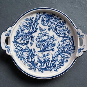 Op de afbeelding: Een rond keramieken serveerblad met twee geïntegreerde handgrepen, voorzien van een witte basis versierd met ingewikkelde blauwe bloem- en rankmotieven. Het ontwerp bevat een centraal bloemmotief omringd door symmetrisch gebladerte.