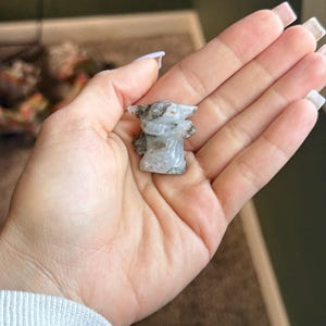 Op de afbeelding: Een klein, gebeeldhouwd beeldje van een personage met grote oren en een gewaad. Het beeldje is gemaakt van een lichtgekleurde steen met grijze en bruine insluitsels. Het beeldje wordt in een open hand gehouden.