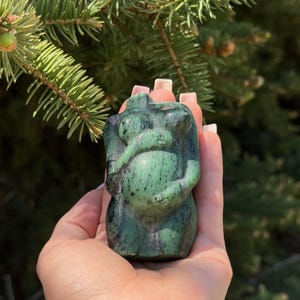 May include: A green and black carved stone sculpture of a pregnant figure holding a baby. The sculpture is held in a hand against a backdrop of green foliage. The stone has a speckled appearance.