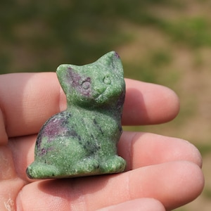 May include: A small, carved stone figurine in the shape of a sitting cat. The sculpture is primarily green with distinct patches of deep magenta and dark grey inclusions throughout the textured surface, depicting ears, eyes, and a nose.
