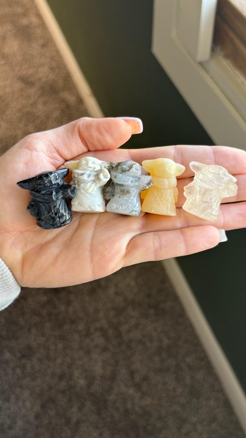 Op de afbeelding: Vijf kleine, gebeeldhouwde beeldjes van een personage met grote oren en een gewaad, in verschillende kleuren, waaronder zwart, wit, grijs, geel en helder. De beeldjes worden in een hand gehouden en tonen de verschillende soorten en kleuren stenen.
