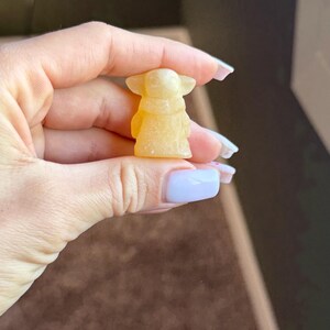 Op de afbeelding: Een kleine, gele figuur van een personage met grote oren en een gewaad. Het beeldje wordt vastgehouden in een hand met verzorgde nagels. De achtergrond is een vage mix van bruin en donkergroen.