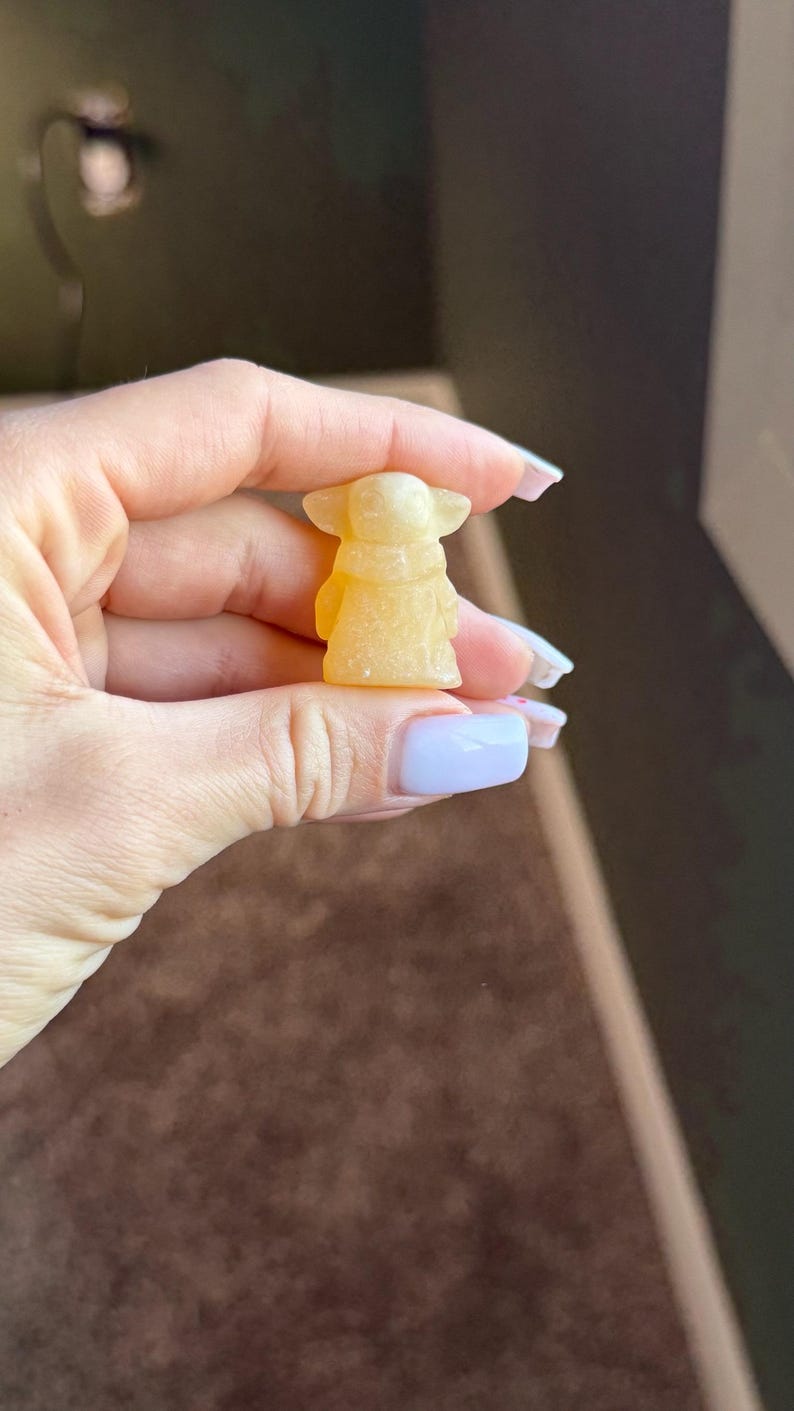 Op de afbeelding: Een kleine, gele figuur van een personage met grote oren en een gewaad. Het beeldje wordt vastgehouden in een hand met verzorgde nagels. De achtergrond is een vage mix van bruin en donkergroen.