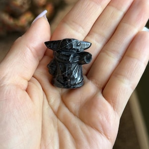 Op de afbeelding: Een klein, zwart beeldje van een personage met grote oren en een gewaad. Het beeldje is gebeeldhouwd uit een donkere steen en wordt in een open hand gehouden. Het beeldje is ongeveer 5 cm hoog.