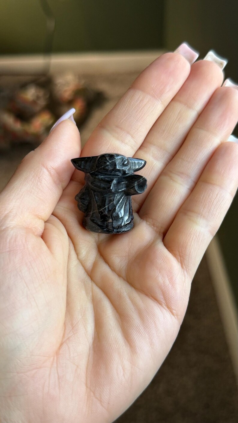 Op de afbeelding: Een klein, zwart beeldje van een personage met grote oren en een gewaad. Het beeldje is gebeeldhouwd uit een donkere steen en wordt in een open hand gehouden. Het beeldje is ongeveer 5 cm hoog.