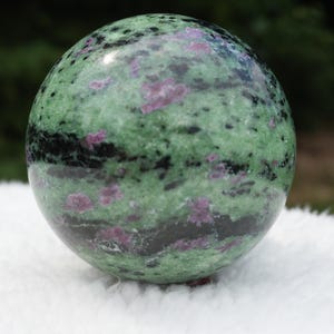 Puede incluir: Una piedra esférica pulida que presenta un patrón moteado de inclusiones minerales en verde claro, verde oscuro y negro, con parches dispersos de color rosa magenta vibrante por toda la superficie lisa y redondeada.