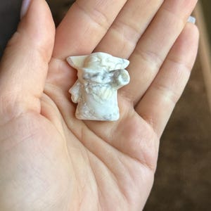 Op de afbeelding: Een klein, wit beeldje van een personage met grote oren en een groot hoofd, vastgehouden in een hand. Het beeldje heeft een textuuroppervlak en is gemaakt van een lichtgekleurd materiaal. Het beeldje is een verzamelobject.