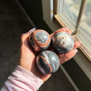 May include: Three spherical decorative stones with a marbled appearance. The stones are primarily dark gray with red and white veining. The stones are held in a hand near a window.