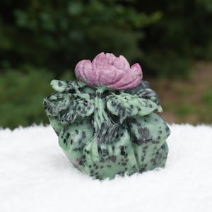 May include: A carved stone sculpture featuring a vibrant pink flower with layered petals resting atop a base of mottled light green and dark black zoisite. The floral carving displays intricate petal details and textured leaves.