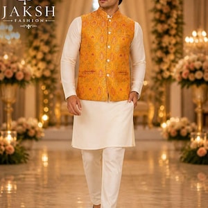 May include: A man in a cream-colored kurta and pants, paired with a yellow patterned vest. The vest has a button-down front. The image features the "JAKSH FASHION" logo. The background is a blurred indoor setting with floral arrangements and warm lighting.