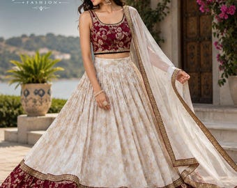 May include: A sleeveless maroon and gold embroidered top and skirt set. The skirt is cream-colored with a gold floral pattern and a maroon floral border. A sheer white scarf with gold trim completes the ensemble. The logo "JAKSH FASHION" is visible.