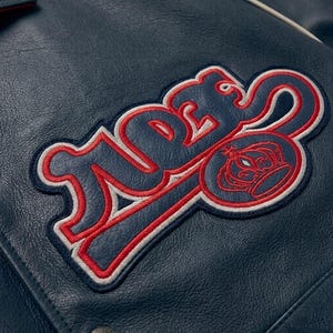 May include: Close-up of a navy blue leather jacket with a large embroidered patch. The patch features the word "TIDES" in a stylized font, outlined in red and white, with a crown emblem.