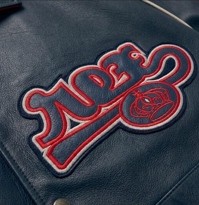 May include: Close-up of a navy blue leather jacket with a large embroidered patch. The patch features the word "TIDES" in a stylized font, outlined in red and white, with a crown emblem.