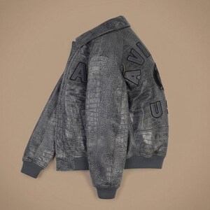 May include: A dark gray bomber jacket with a textured crocodile skin pattern. The jacket features a classic collar, ribbed cuffs and hem, and large black block letters spelling AVIATOR across the back, along with a circular emblem patch.