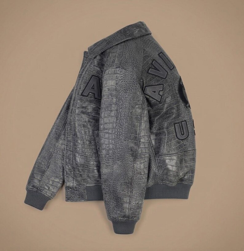 May include: A dark gray bomber jacket with a textured crocodile skin pattern. The jacket features a classic collar, ribbed cuffs and hem, and large black block letters spelling AVIATOR across the back, along with a circular emblem patch.