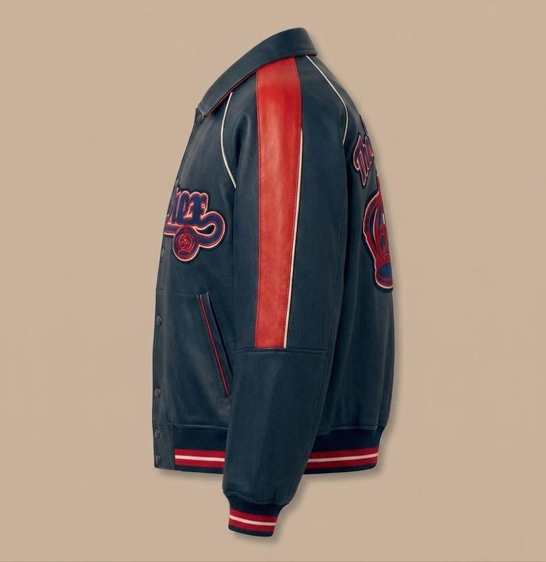 May include: A navy blue varsity jacket with red and white accents. The jacket features a red stripe down the sleeve and patches with text. The cuffs and hem have red and white striped detailing.