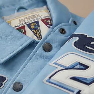Puede incluir: Una chaqueta universitaria de cuero azul claro con letras y parches bordados en azul oscuro y blanco. La etiqueta interior dice AVIREX AUTHENTIC VARSITY SPORTS EQUIPMENT. La chaqueta incluye botones de presi&oacute;n de metal oscuro grabados con AVIREX.