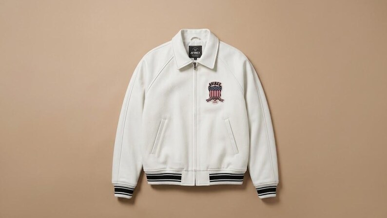 May include: A white zip-up jacket featuring a fold-down collar, long sleeves, and two front pockets. The chest displays an embroidered patch with the text AVIREX above a shield graphic featuring red and white stripes and blue stars.