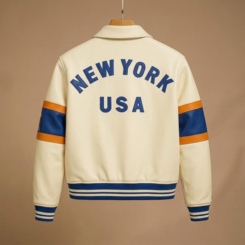 May include: A cream-colored varsity jacket featuring blue text on the back that reads NEW YORK USA. The sleeves have blue and orange horizontal stripes, and the cuffs and hem are finished with blue and white ribbed knit trim on a hanger.