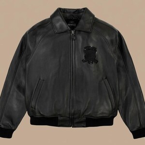 May include: Black leather jacket with a collared neckline, a zipper closure, and a patch on the chest. The jacket has two front pockets and ribbed cuffs and hem. The brand name "AVIREX" is visible on the collar.