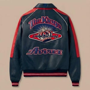 May include: A navy blue leather jacket with red accents on the sleeves and collar. The back features an embroidered design with the text "The Kings", "Gold Kickoff", and "Avirex". The jacket has a ribbed hem and cuffs with red and white stripes.