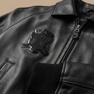 May include: Close-up of a black leather jacket with a zipper closure and a patch featuring an emblem. The jacket has a ribbed cuff and a collared neckline. The emblem is a stylized design with wings and text.