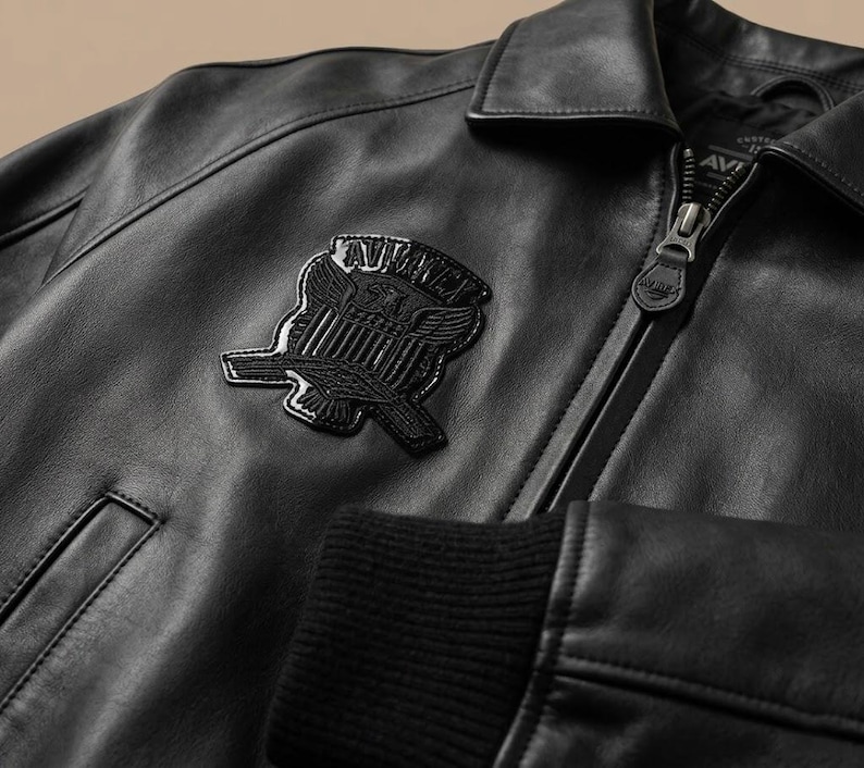 May include: Close-up of a black leather jacket with a zipper closure and a patch featuring an emblem. The jacket has a ribbed cuff and a collared neckline. The emblem is a stylized design with wings and text.